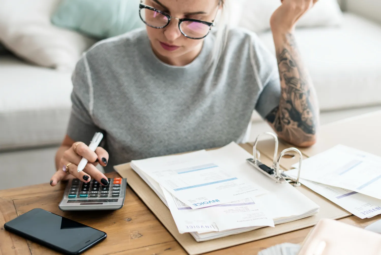Mujer con gafas usando una calculadora mientras revisa documentos y facturas en una mesa de trabajo.